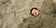 Ein Junge hat sich in Sand eingebuddelt, nur noch der Kopf ragt raus.