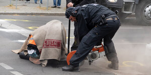 Frau mit Wolldecke miut Aufschrift "Polizei" über dem Kopf neben einem Polizisten mit Kreissäge