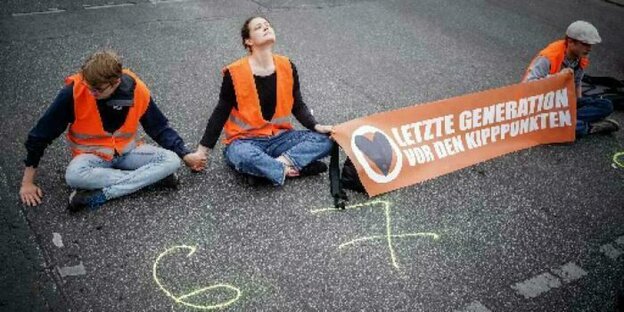 Klimaaktivisten der Lezten Generation sitzen auf einer Straße, vor sich sind die Ziffern 6 und 7 mit Kreide auf den Asphalt gemalt