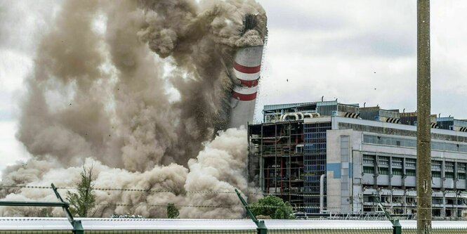 Ein großer Schornstein fällt in einer großen Rauchwolke in sich zusammen, davor ein industriell aussehendes Gebäude.