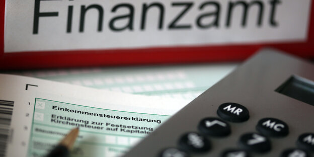 Ein Vordruck für Einkommenssteuererklärung liegt neben einem Aktenordner mit der Aufschrift Finanzamt, einem Stift und einem Taschenrechner.