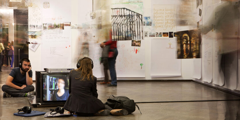 Ein Raum mit einer Kunstinstallation. An der Wand hängen Bilder. Auf dem Boden stehen zwei Fernseher, vor denen zwei Menschen mit Kopfhörern sitzen. Andere Menschen bewegen sich durch den Raum.