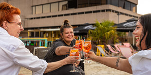 Drei Frauen prosten sich mit Aperol Spritz in einer Strandbar zu, im Hintergrund ist ein Hochhausbau erkennbar.