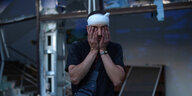 Ein Mann trägt einen Verband um den Kopf und hält sich mit beiden Hönden die Augen zu. Im Hintergrund sind Trümmer und ein beschädigtes Gebäude zu erkennen