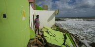 Ein Mann steht in den Trümmern seines grün gestrichenen Hauses und schaut auf das Meer, dessen Wellen direkt vor seinem Haus brechen