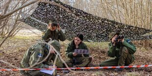 Kinder in Militäruniform unter einem Tarnnetz schauen druch Ferngläser, sitzend auf dem Boden