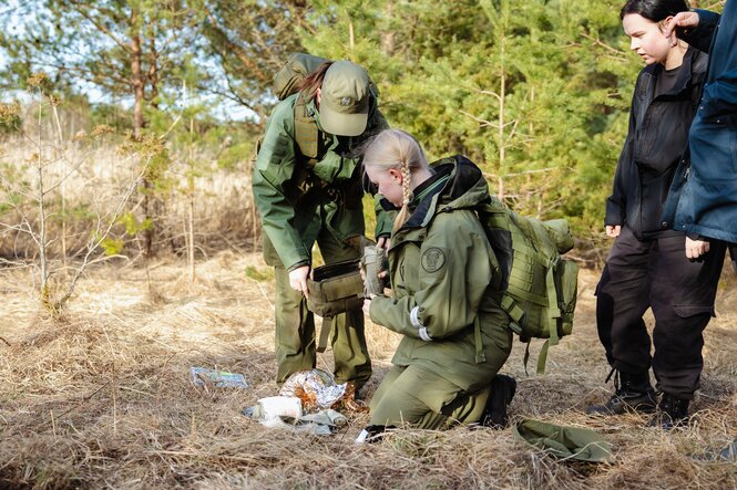 Kinder in grüner Uniform, ein Mädchen mit blonden Zöpfen kniet auf dem Boden und packt schmutziges Verbandszeug in eine Tasche, die ihr entgegengehalten wird.