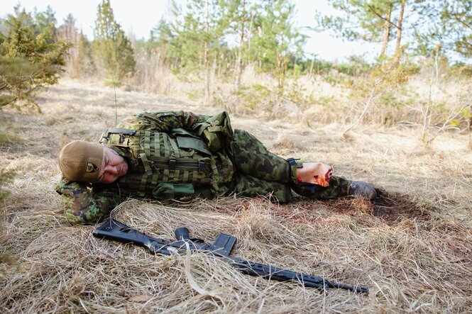 Ein Mann in Uniform liegt im Gestrüpp, ein Bein ist aufgerissen, eine Attrappe, neben ihm ein Maschinengewehr