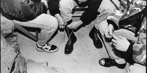 Drei junge Männer sitzen auf einer Bank, einer schnürt sich die Springerstiefel, der junge Mann neben ihm trägt Stahlkappenschuhe