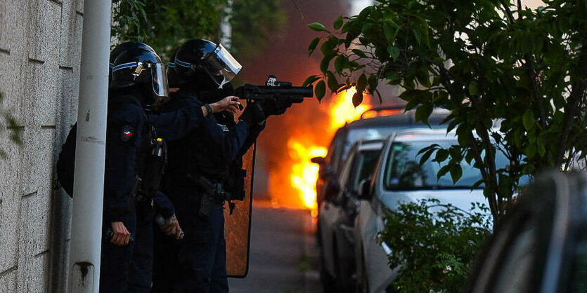 Zwei Polizisten am Strassenrand mit einer Waffe, dahinter sieht man Flammen