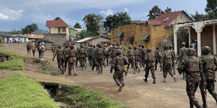 Soldaten laufen auf einer unbefestigten Straße, Häuser einer Kleinstadt im Hintergrund