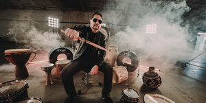 Dave Lombardo, umgeben von Trommeln und Trockeisnebel, reckt einen Drumstick in die Kamera