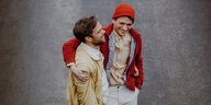 Die Musiker Marco Mlynek und Max Andrzejewski laufen Arm in Arm über einen Untergrund aus Asphalt. Mlynek trägt einen Bart und eine gelbe Baumwolljacke. Andrzejewski trägt eine orange-rote Mütze und eine rote Jacke. Sie schauen im Moment versunken von der Kamera weg und lachen.
