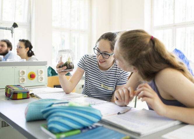 Zwei Schülerinnen schauen sich im Klassenraum eine Bodenprobe im Glas an