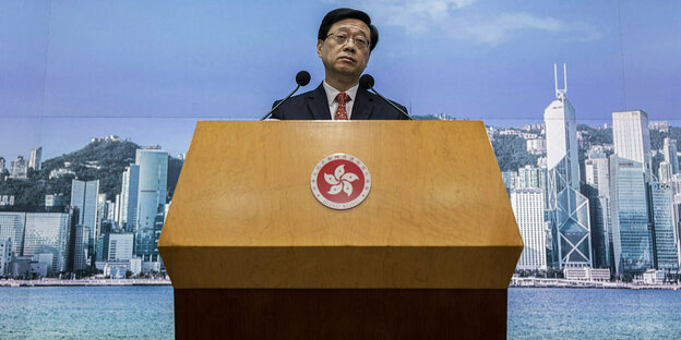 John lee steht an einem Rednerpult, im Hintergrund ist die Stadt Hongkong zu sehen