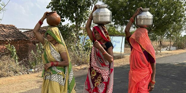 Drei indische Frauen gehen auf einer Dorfstraße und tragen Wasserbehälter auf ihrem Kopf
