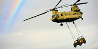 Ein gelber Transporthuschrauber transportiert einen Geländewagen, der mit Seilen am Hubschrauber hängt
