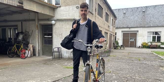 Ein Mann steht in Wildlederjacke und schwarz-weiß-gestreiftem T-Shirt mit Fahrrad und einer Tasche auf dem Rücken auf einem Hof.