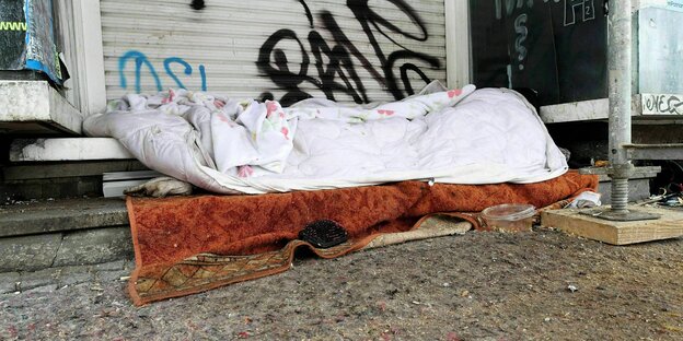 Schlafplatz eines Obdachlosen zwischen Müll, Unrat, alten Veranstaltungsplakaten und einem Baugerüst am Straßenrand vor einem sanierungsbedürftigem Mehrfamilienhaus.