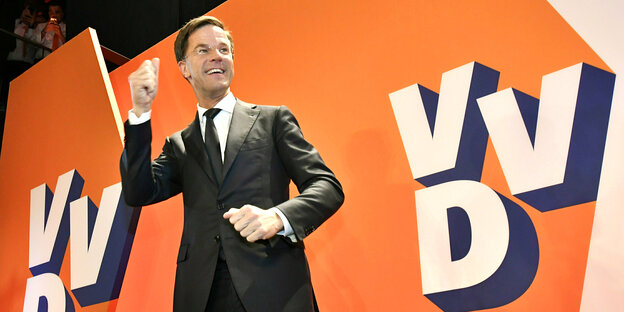 Mark Rutte steht lächelnd vor einer orangefarbenen Wand mit dem Logo der VVD