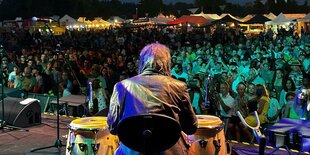 Wir sehen den rücken eines Mannes der auf Congas trommelt, im Hintergrund das Publikum des Sunrise Festivals von bunten Lichtern beleuchtet