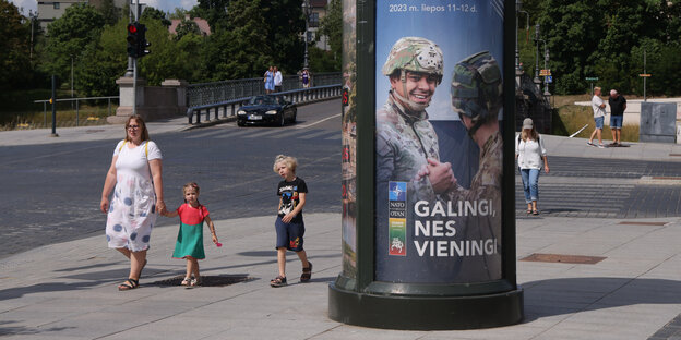 Eine Werbesäule mit Soldaten abgebildet, daneben läuft eine Frau mit zwei Kindern vorbei