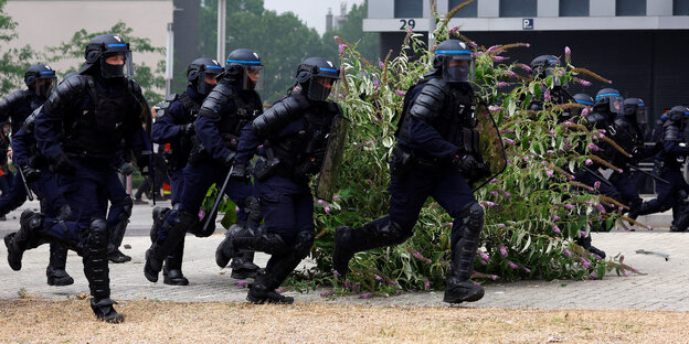 Eine Gruppe französischer Polizist*innen in Riot Gear rennt von links nach rechts durch das Bild