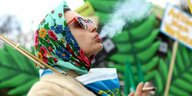 Eine Frau mit buntem Kopftuch raucht vor einem großen Plastik-Cannabisblatt genüsslich einen Joint