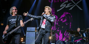 Links ein Mann am Bass, rechts Sänger Billy Idol in schwarzer Lederjacke
