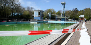 Rot-weißes Absperrband ist vor einem Freibadbecken gespannt. Im Hintergrund ist ein Sprungturm zu sehen.