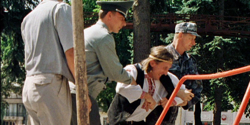 Ein Mann in belarussischer Tracht wird abgeführt, im Vordergrund eine Schubkarre Dung mit einem Bild von Alexander Lukaschenko