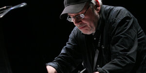 Der Musiker Wolfgang Seidel steht auf einer Bühne, vor ihm steht ein Laptop. Er trägt ein schwarzes Hemd und eine schwarze Baseball-Cap.