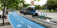 Ein blauer Fahrradweg führt an Moped-Parkplätzen und einer Autostraße vorbei