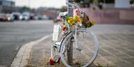Ein sogenanntes Geisterrad, also ein weiß angemaltes fahrrad, löehnt an einer Laterne. Am Lenker sind Blumen befestigt.
