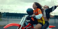 Zwei junge Frauen fahren auf einem Motorrad, die Haare flattern im Wind, die Frau auf dem Sozius breitet die Arme aus