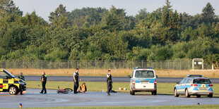 Menschen sitzen auf einem Rollfeld, daneben Polizisten und Autos