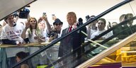 Trump fährt eine Rolltreppe runter, im Hintergrund Fans mit T-Shirts, auf denen "Trump - Make America Great Again" steht.