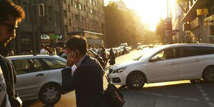 Mann mit Smartphone am Ohr und Aktentasche unterm Arm läuft im über die Kreuzung am Checkpoint Charlie, gleichzeitig biegen zwei Autos ab