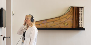 Eine blonde Frau hat einen Kopfhörer auf, im Hintergrund hängt ein altmodisches tasrninstrument an der Wand