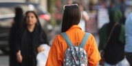 Eine Frau von hinten fotografiert, hat ihr langes dunkles Haar offen zu einer Seite gelegt und eine Sonnebrille ins Haar gesteckt, sie trägt eine orange Jacke und einen Rucksackkt