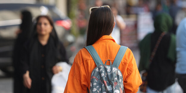 Eine Frau von hinten fotografiert, hat ihr langes dunkles Haar offen zu einer Seite gelegt und eine Sonnebrille ins Haar gesteckt, sie trägt eine orange Jacke und einen Rucksackkt