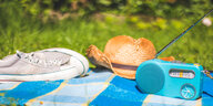 türkisfarbenes kleines Radio auf einer Picknickdecke, daneben Sonnenhut und Turnschuhe