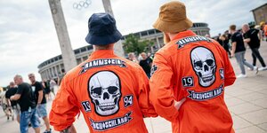 Im Hintergrund ist das Berliner Olympiastadion zu sehen. Im Vordergrund sehen wir die Rücken von zwei Männern in orangefarbenen Overalls, darauf jeweils ein Totenkopf und die Aufschrift: "Rammstein, Deutschland, 1994"