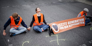 Aktivist*innen der letzten Generation sitzen mit einem Banner auf der Straße, einer hat die Hand aufgeklebt