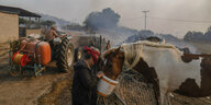 Eine Frau mit Mundschutz gibt einem Pferd Wasser in einem Eimer, ein Mann mit Traktor spritzt Löschwasser auf die brennende Weide