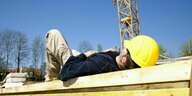 Ein Mann mit gelbem Schutzhelm liegt auf einem Holzträger auf einer Baustelle und macht ein Nickerchen