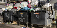 Drei schwarze Mülltonnen, in denen sich Müllsäcke stapeln, stehen vor einer Wand. Auch vor den Müllonnen liegt Dreck und Müll.