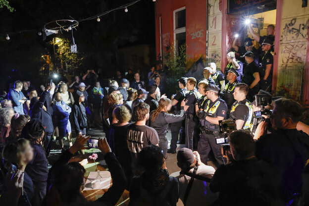 Polizisten stehen vor dem Eingang eines Hauses, davor protestieren Menschen