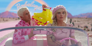 Barbie rechts, gespielt von Margot Robbie, fährt ein rosa Auto. Ken, gespielt von Ryan Gosling, sitzt daneben und hält knall-gelbe Rollschuhe in die Höhe