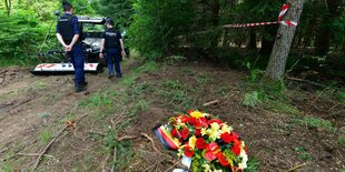 Ein Totenkranz aus Blumen liegt in einem Waldstück, im Hintergrund Männer in blauen Uniformen